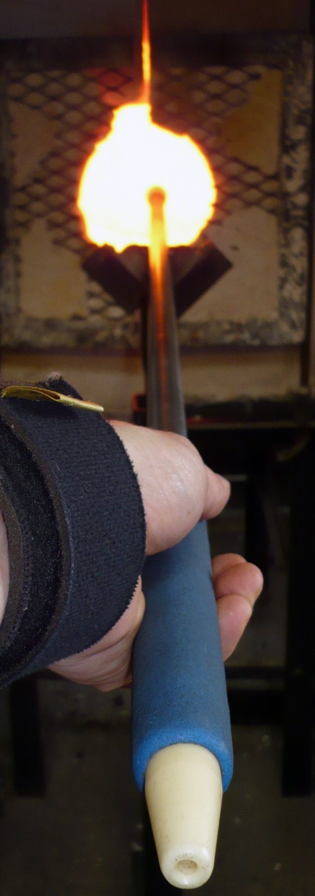 Pre-Heating the BlowPipe in the Glory Hole A hand holding a tool with a flame at the tip, used in glassblowing.